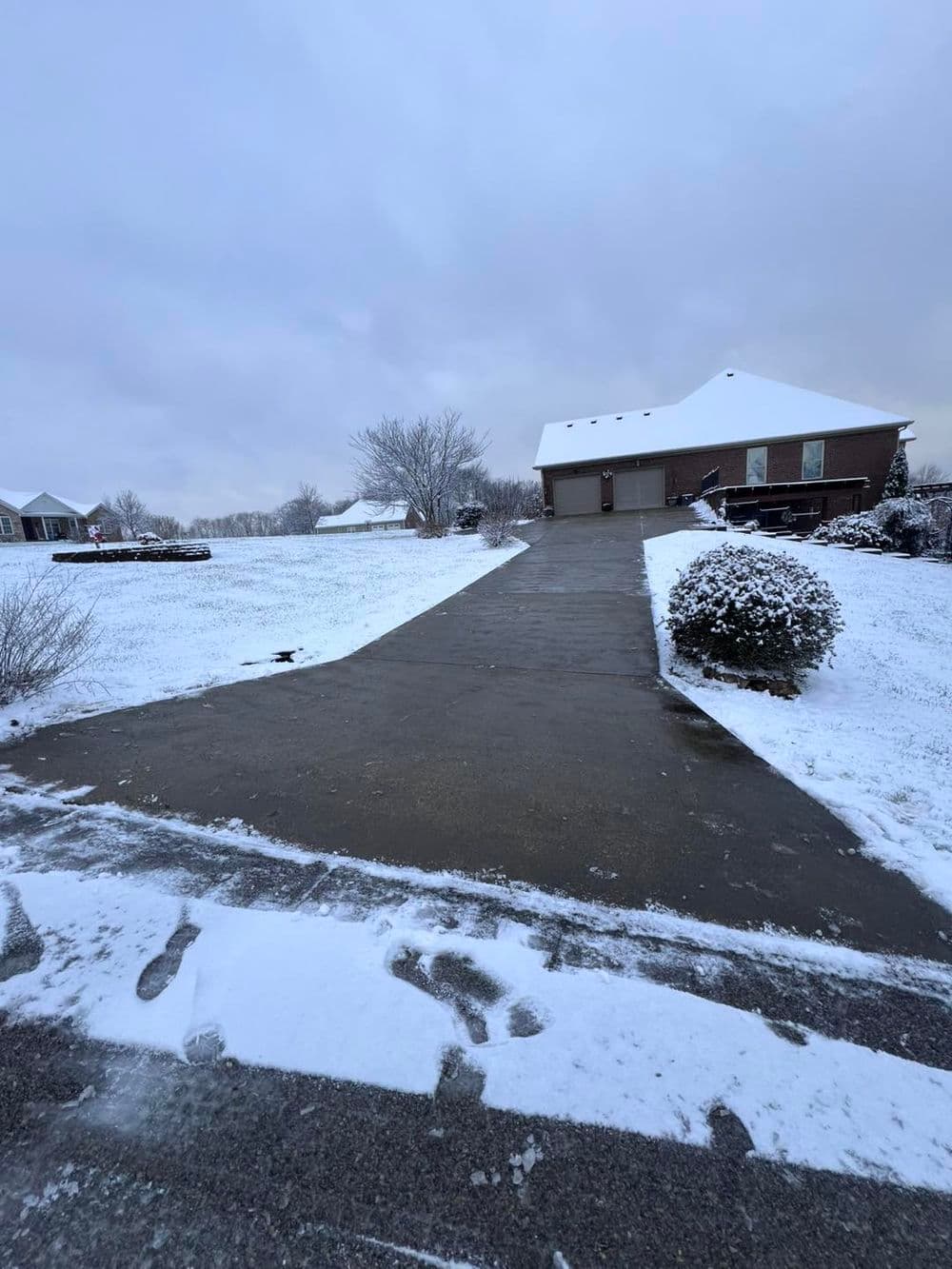 Snow-covered driveway leading to a house in a winter landscape under a cloudy sky.