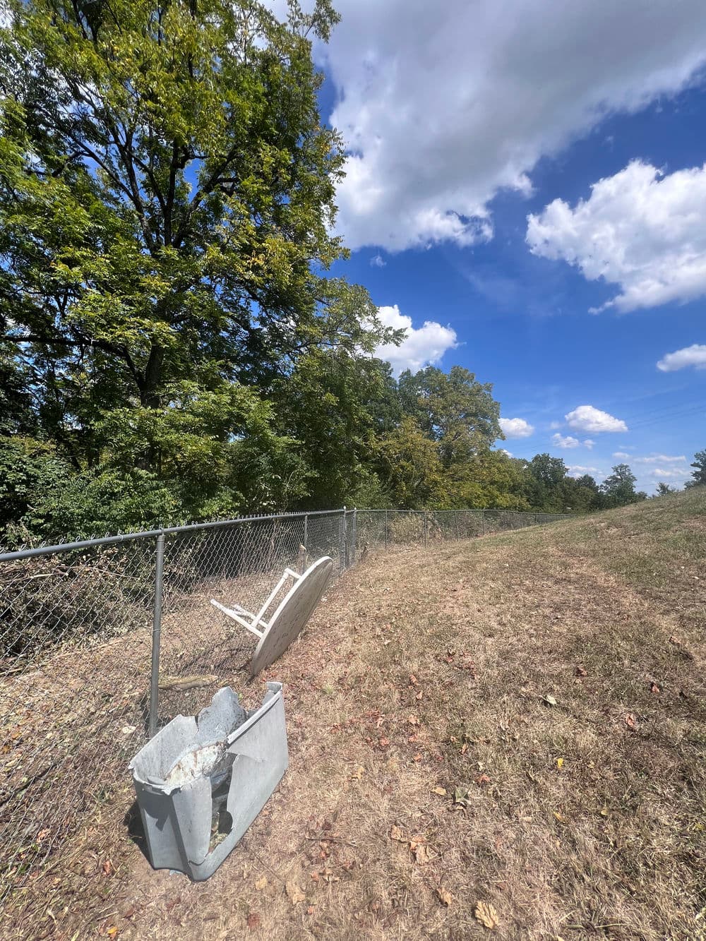 Discarded white chair and trash bin next to a fenced field under a blue sky with clouds.