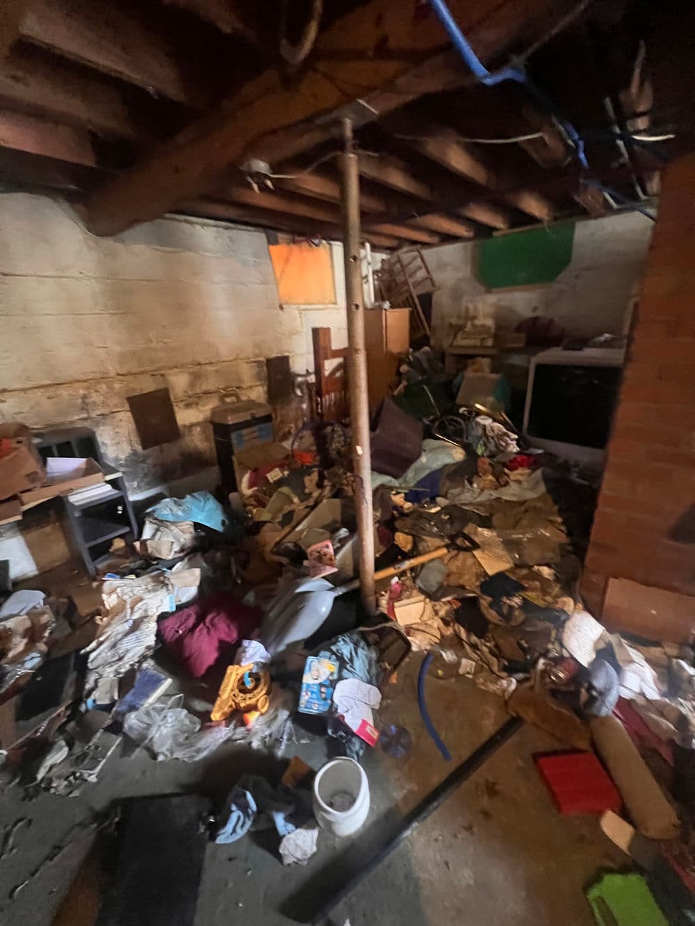 Cluttered basement filled with piles of discarded items, boxes, and debris under wooden beams.