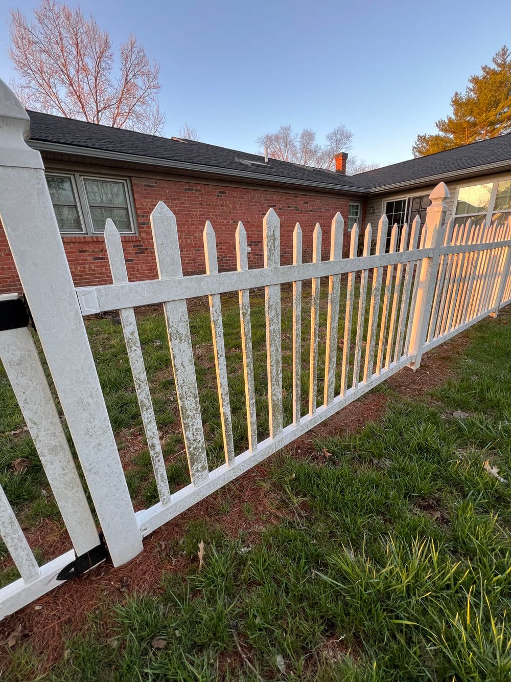 White picket fence in front of a brick house with green grass and evening light.
